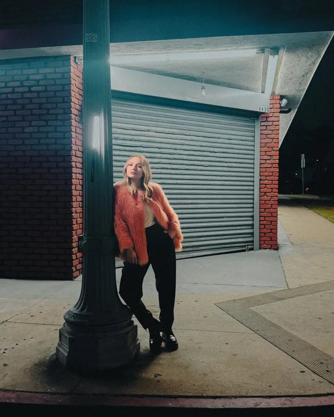 Hayden Panettiere wearing an orange faux fur coat leans against a lamppost at night.