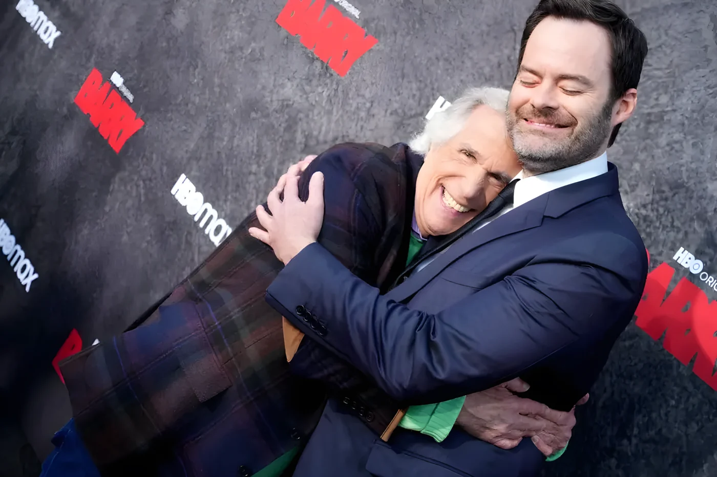 Henry Winkler, in a plaid jacket, hugs Bill Hader, in a navy suit, as both laugh in front of a Barry and HBO Max backdrop.