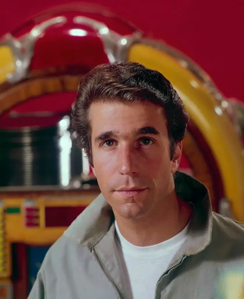 Henry Winkler, with dark swept-back hair and a grey zip jacket over a white T-shirt, looks at the camera in front of a colourful vintage jukebox against a red wall.