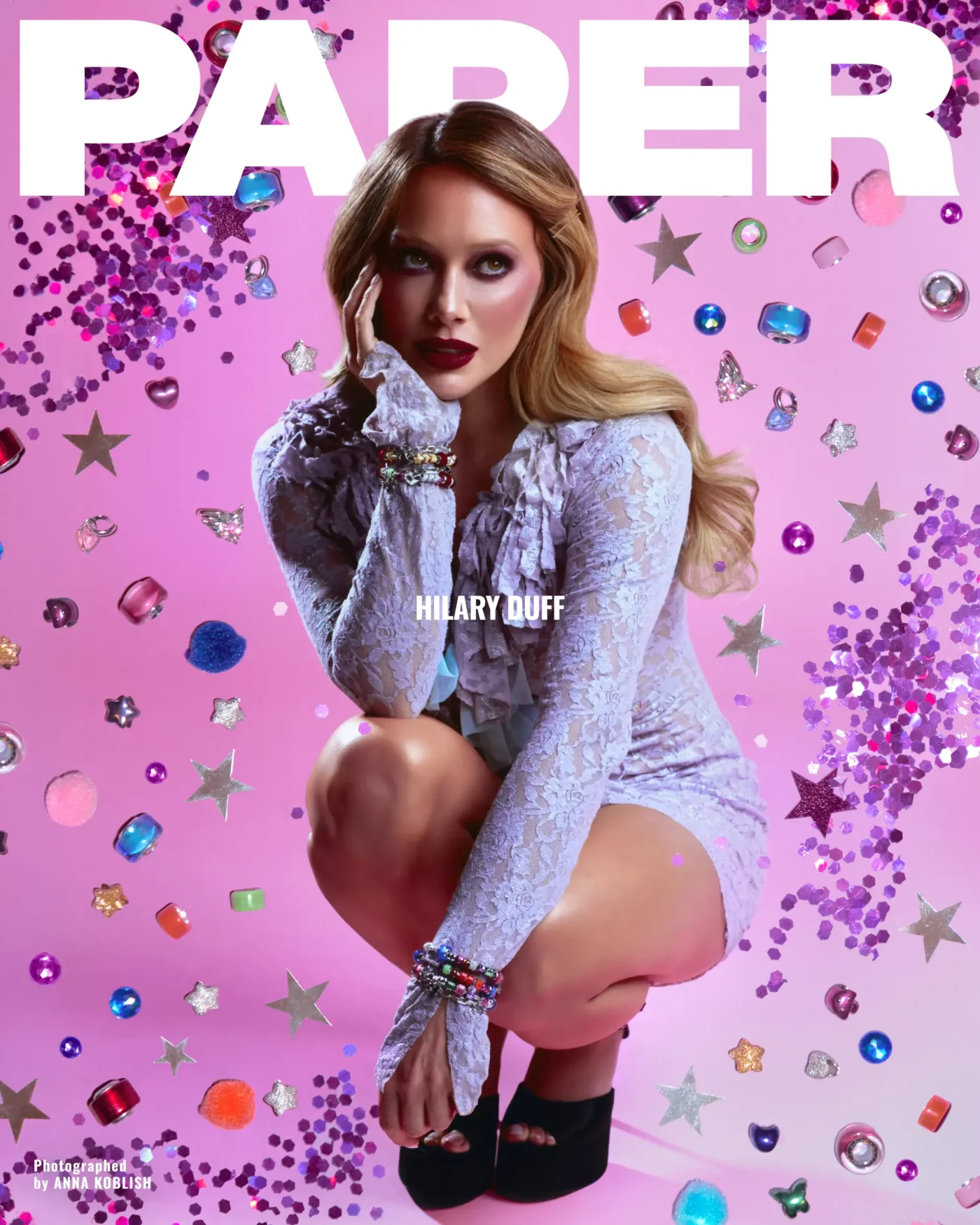 Hilary Duff crouches in a lavender lace dress against a pink background filled with floating stars, beads, and glitter, for Paper magazine.