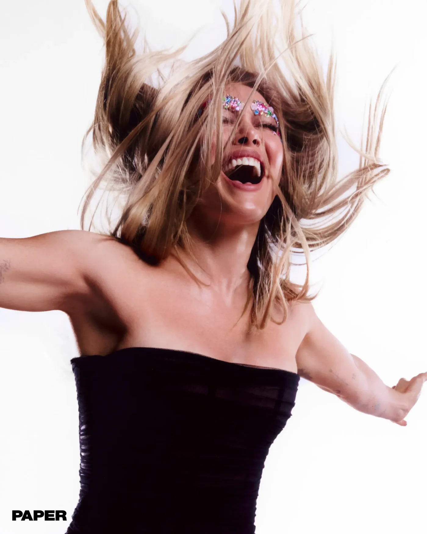Hilary Duff laughing with her head thrown back and blonde hair flying, wearing a black strapless top against a white background.