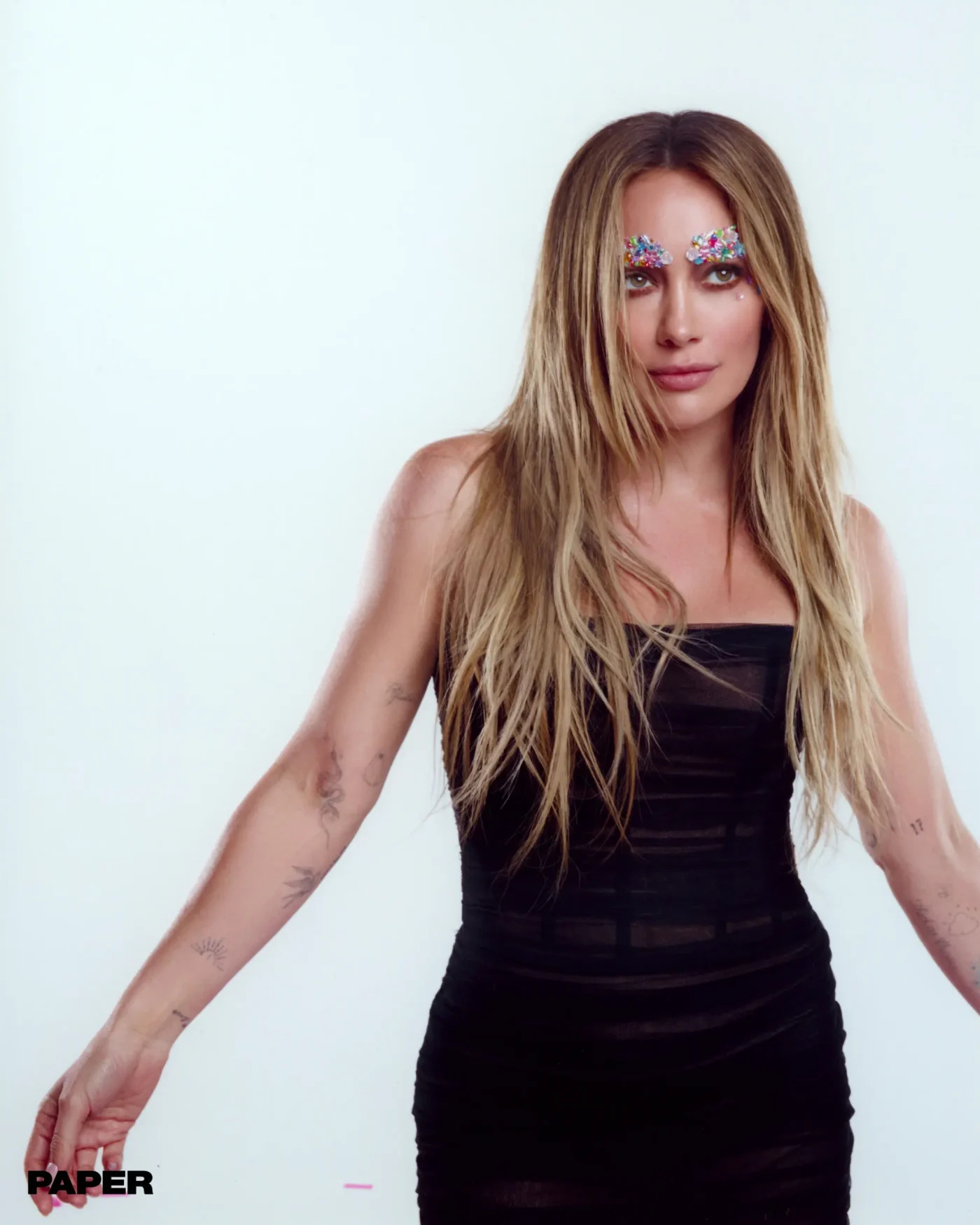 Hilary Duff posing in a black strapless dress with colorful gemstones above her eyes.