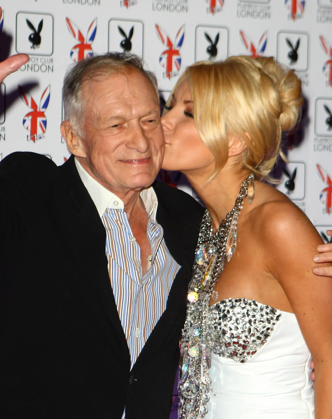 Hugh Hefner and Crystal Harris posing together in front of a backdrop with Playboy Club London logos.