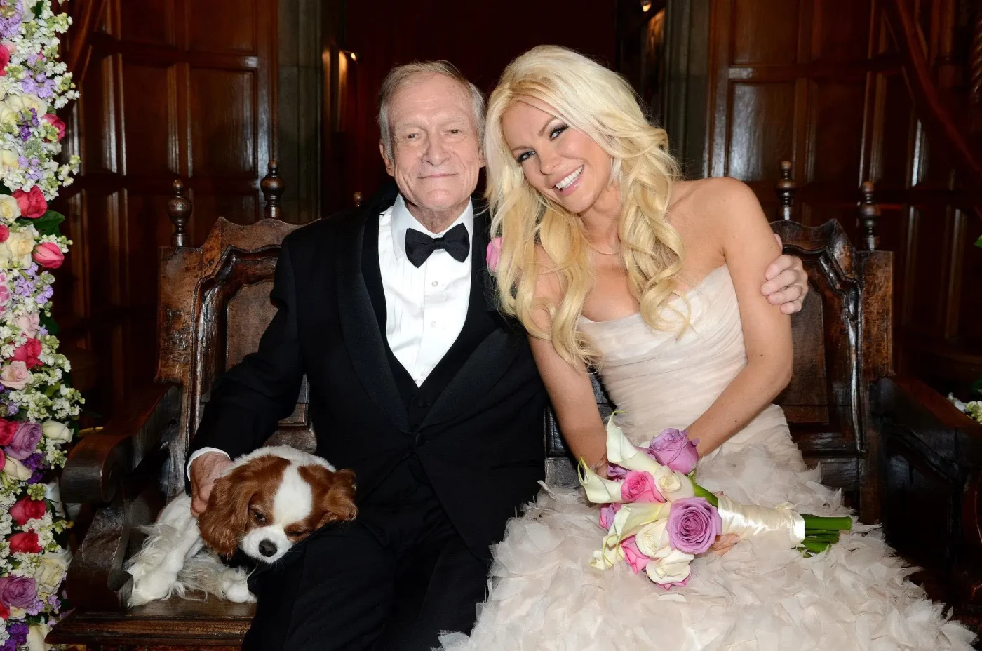 Hugh Hefner and Crystal Harris sitting together on a wooden bench with a small dog and a bouquet of flowers.