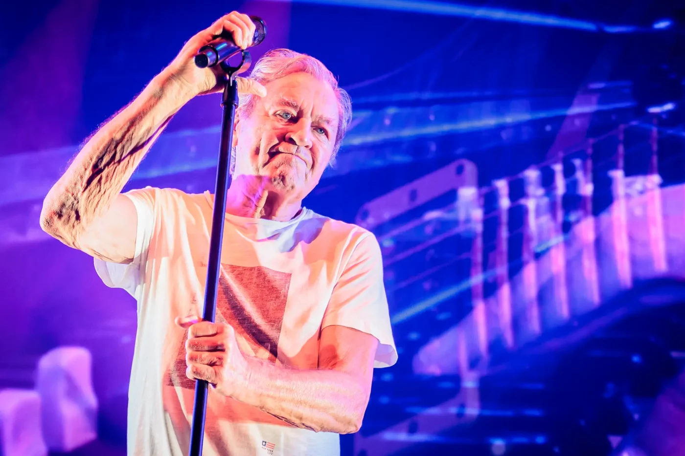 Ian Gillan of Deep Purple performs on stage, holding a microphone stand under blue lighting.