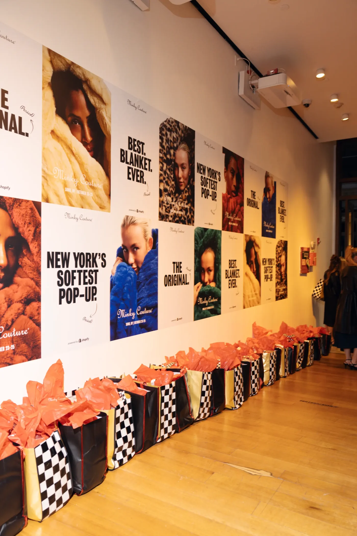 Interior view of a Minky Couture pop-up shop featuring a wall covered in promotional posters and a line of checkered shopping bags filled with orange tissue paper along the floor.