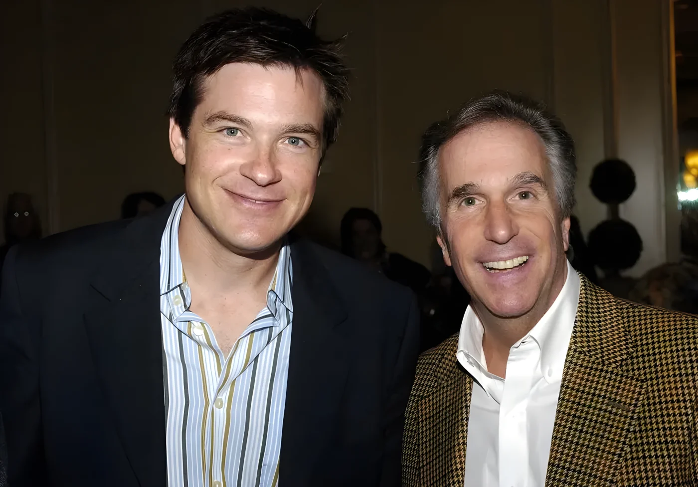 Jason Bateman, in a navy blazer and striped open-collar shirt, smiles beside Henry Winkler, who wears a houndstooth jacket over a white shirt, at an indoor luncheon.