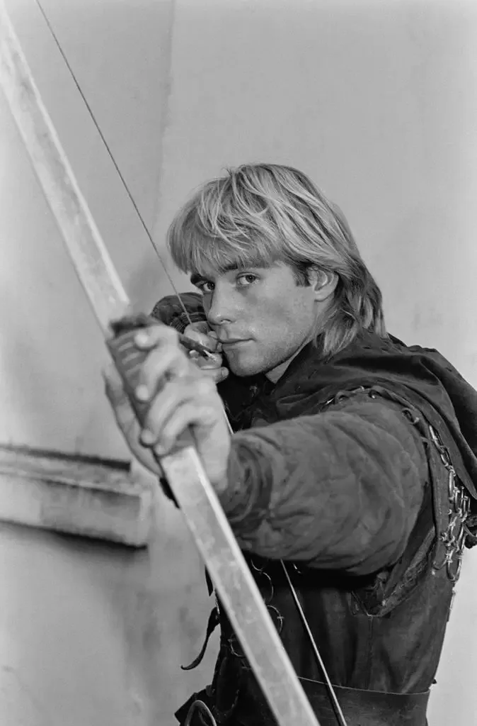 Jason Connery, with long blonde hair, aiming a bow and arrow.