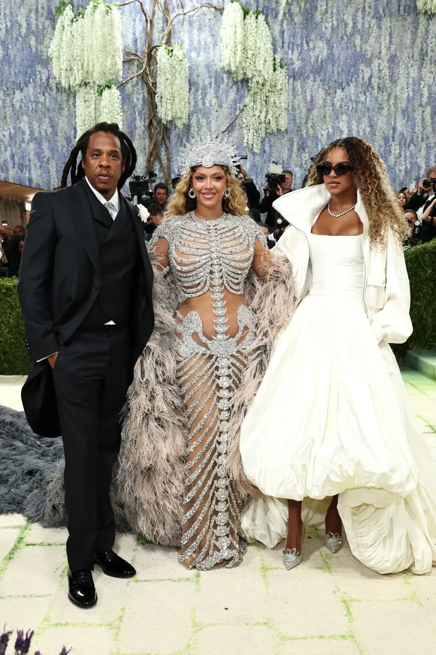 Jay-Z, Beyoncé, and Blue Ivy Carter pose side by side at the Met Gala. Jay-Z is wearing a black suit and patent leather loafers; Beyoncé is wearing a see-through, skeleton-inspired evening gown in silver with a matching crown; and Carter is wearing a white dress with a voluminous high-low skirt and a matching bomber jacket.