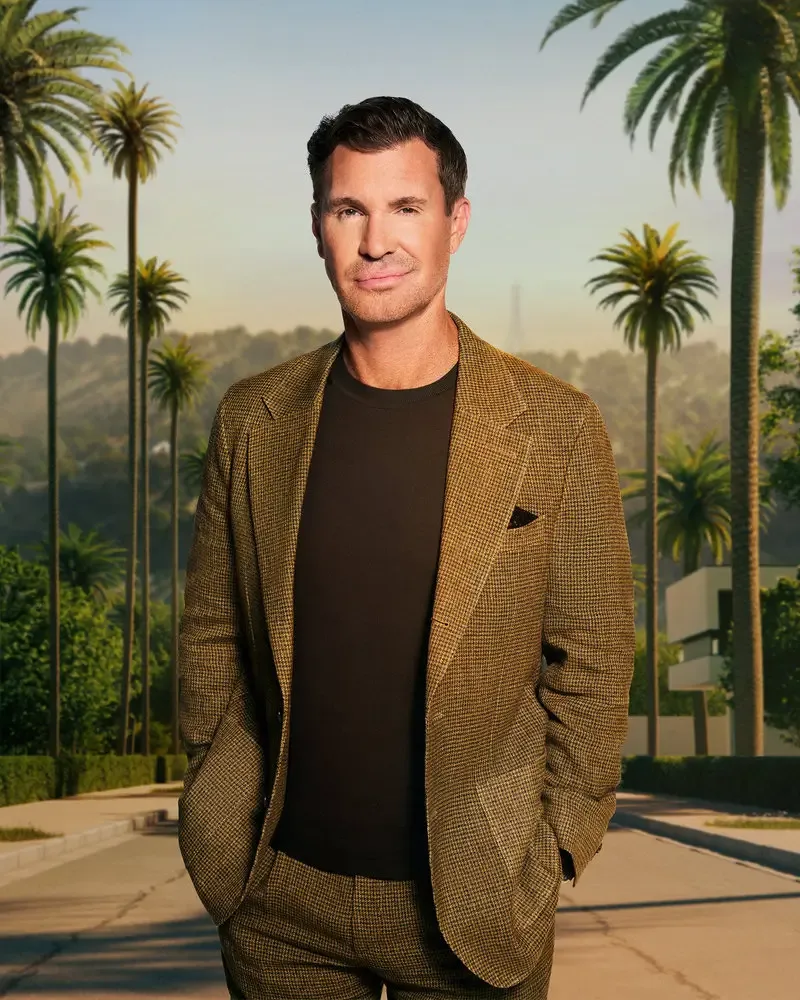 Jeff Lewis in a brown patterned suit and black t-shirt standing outside with palm trees.