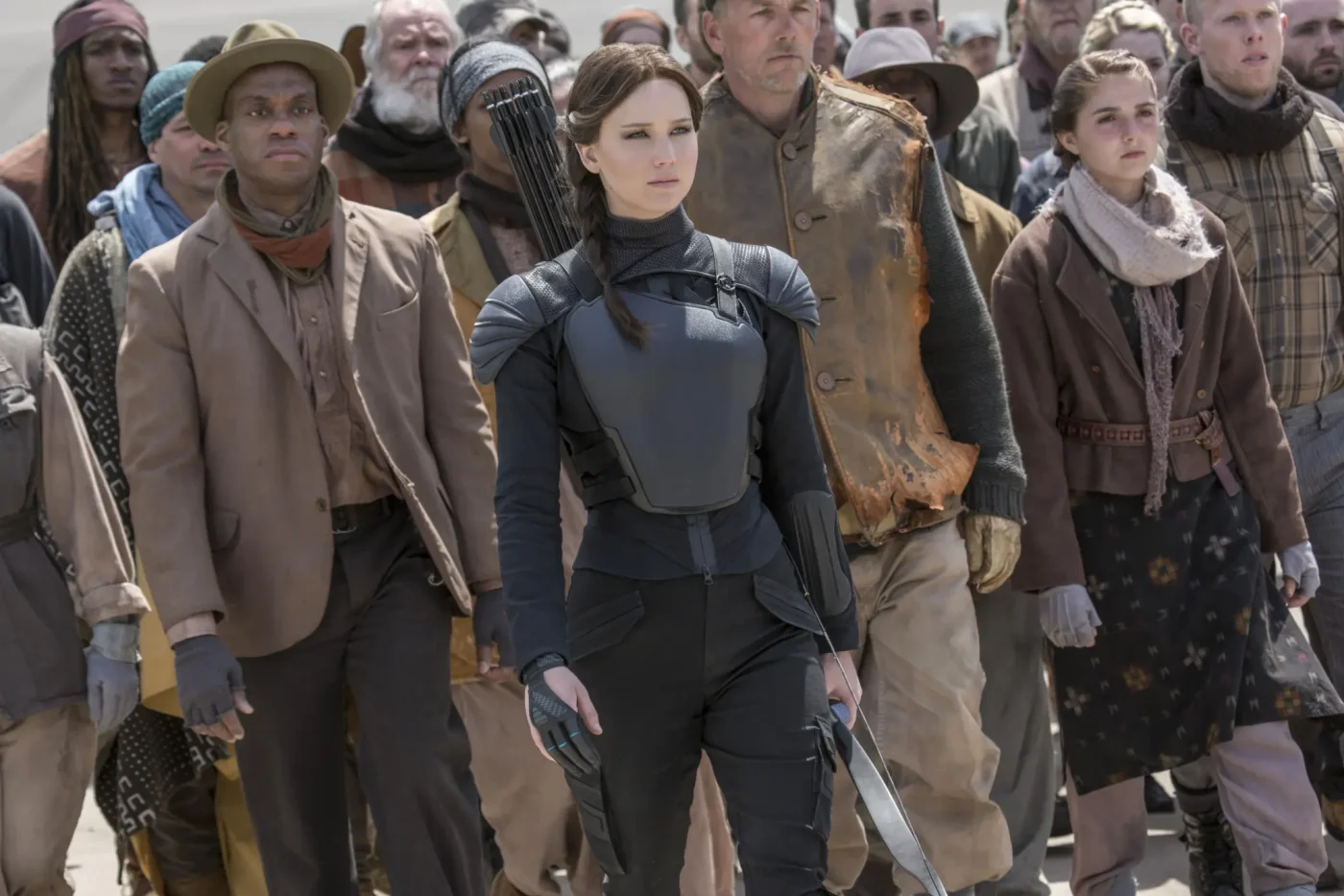 Jennifer Lawrence as Katniss Everdeen wearing black tactical gear and carrying a bow and arrow, surrounded by a crowd in The Hunger Games: Mockingjay - Part 2.