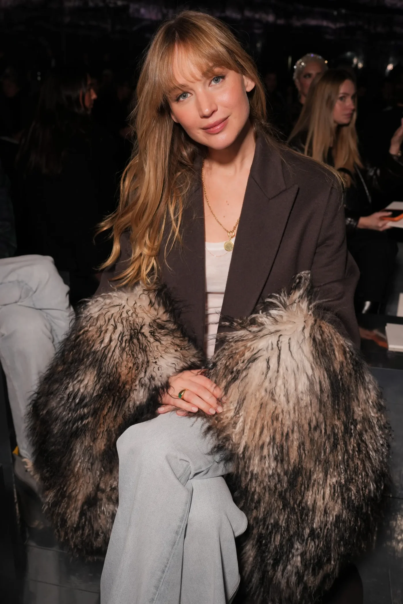 Jennifer Lawrence seated in the front row at a Dior fashion show wearing a blazer with fur cuffs.