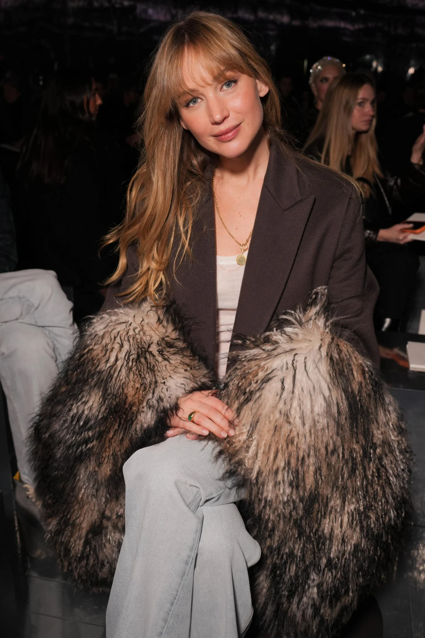 Jennifer Lawrence sitting in the front row at a fashion show, wearing a dark blazer with furry cuffs and light-colored pants.