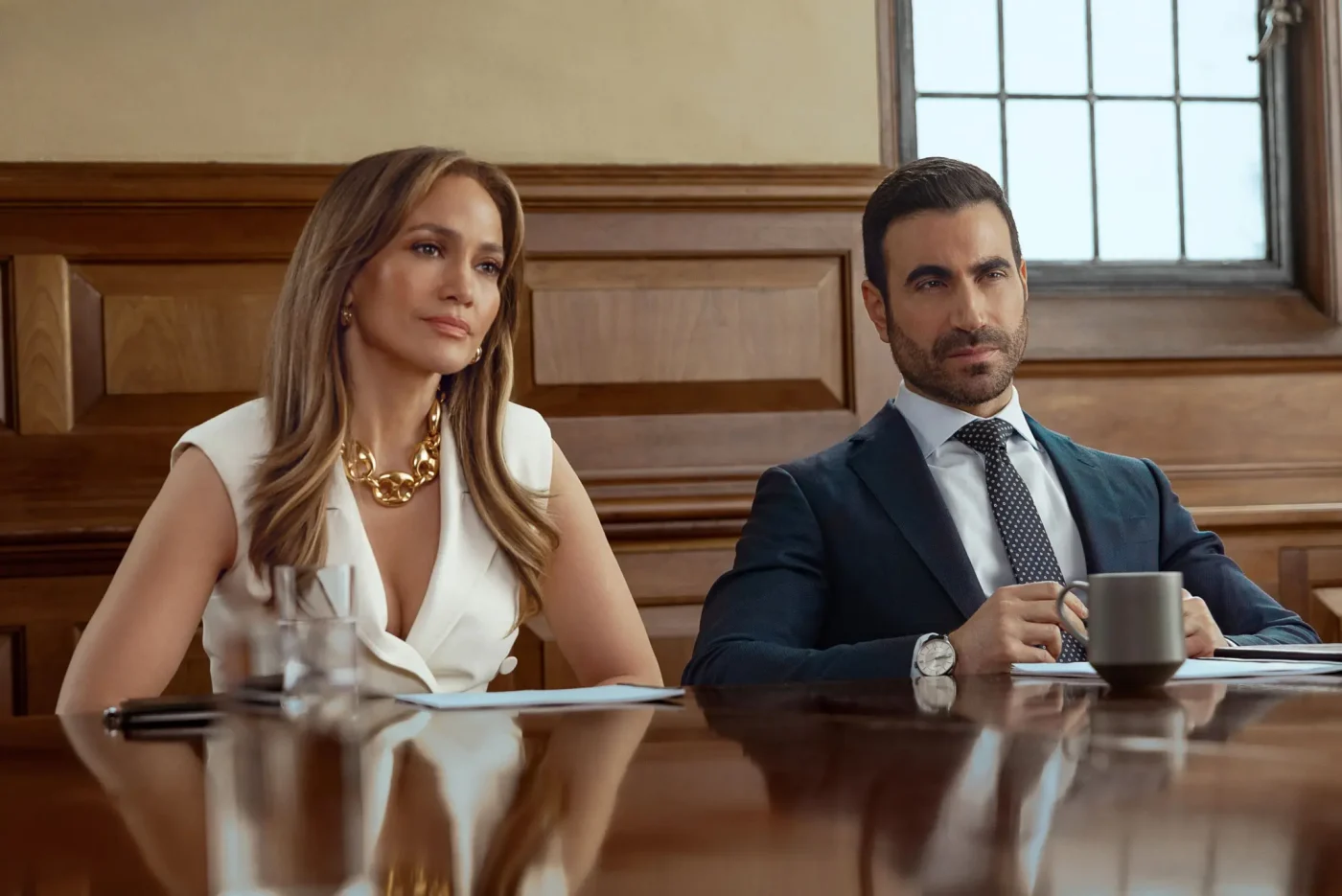 Jennifer Lopez and Brett Goldstein seated at a wooden conference table in Office Romance.
