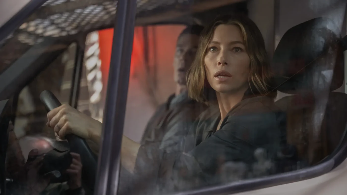Jessica Biel driving a van with a worried expression while looking ahead, with a male passenger visible in the background.
