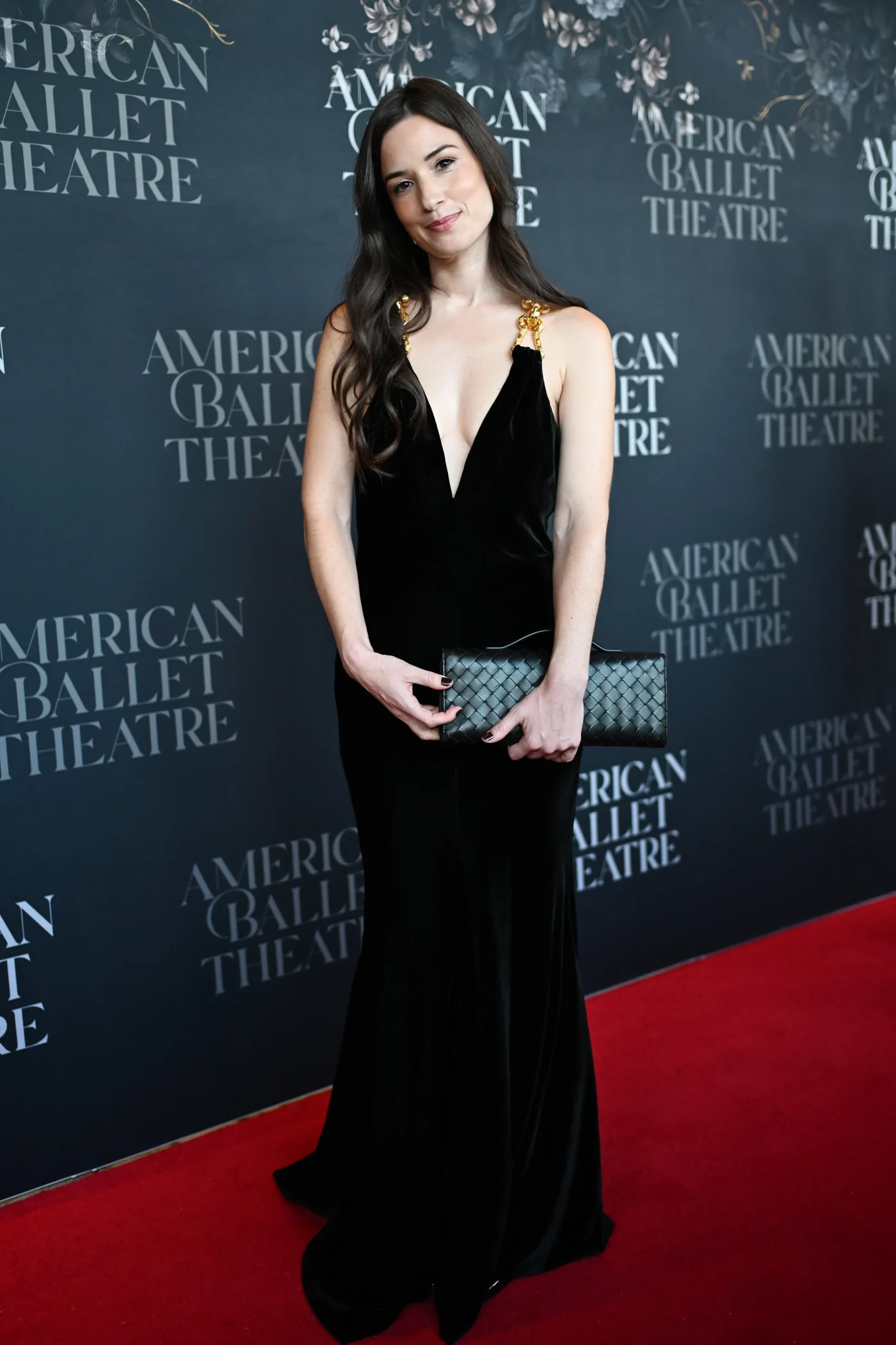 Jessica Whitaker arrives at the American Ballet Theatre Fall Gala, wearing a black dress and holding a clutch, in front of a branded backdrop.