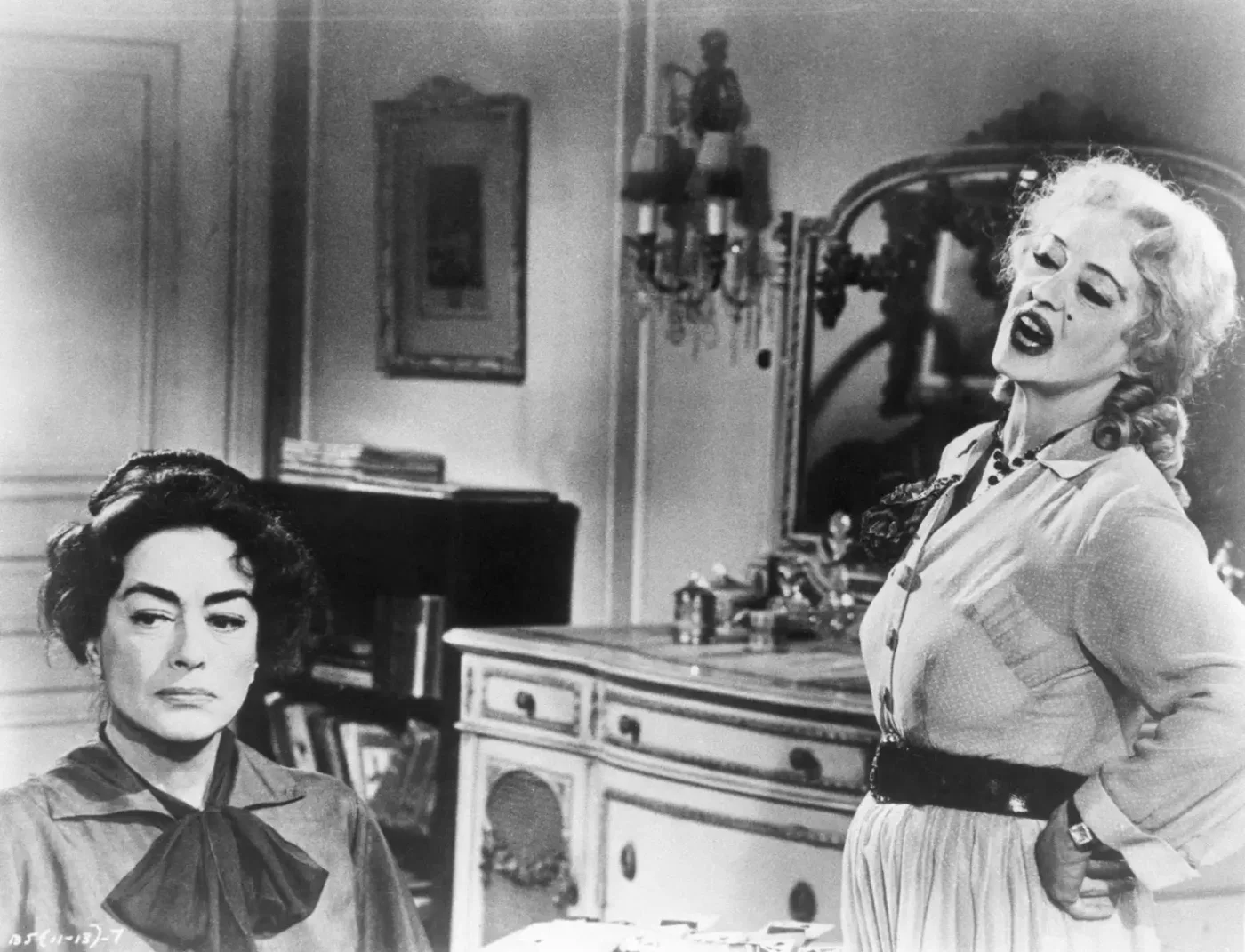 Joan Crawford seated with an unimpressed expression while Bette Davis sings dramatically in a domestic setting
