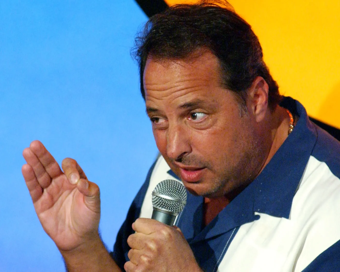 Jon Lovitz holding a microphone and gesturing while speaking on stage.