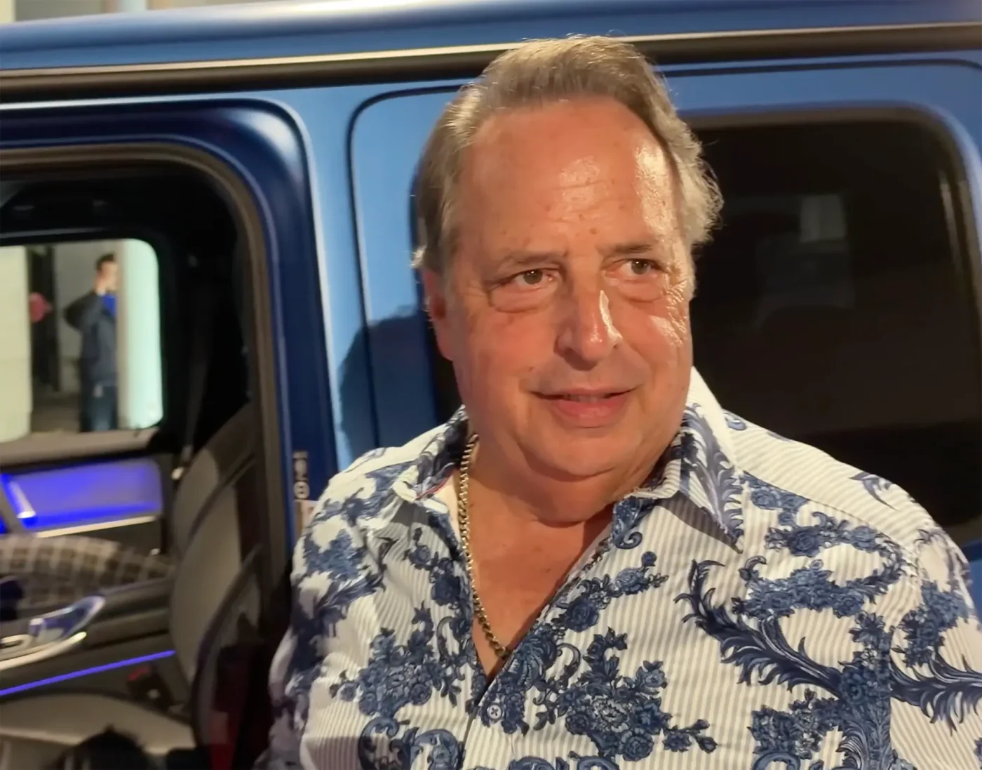 Jon Lovitz standing in front of a blue car.