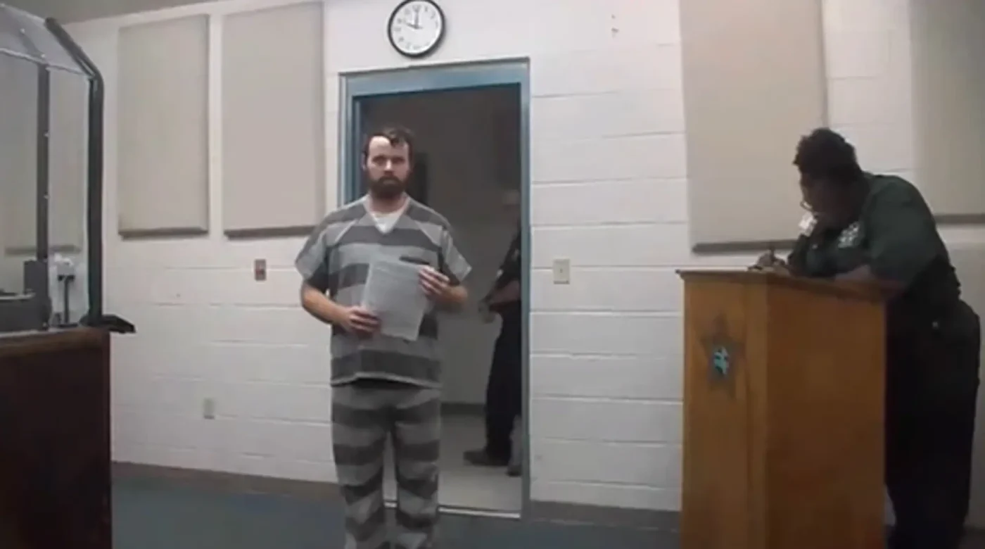 Joseph Duggar standing in a courtroom in a striped prison uniform holding a paper.