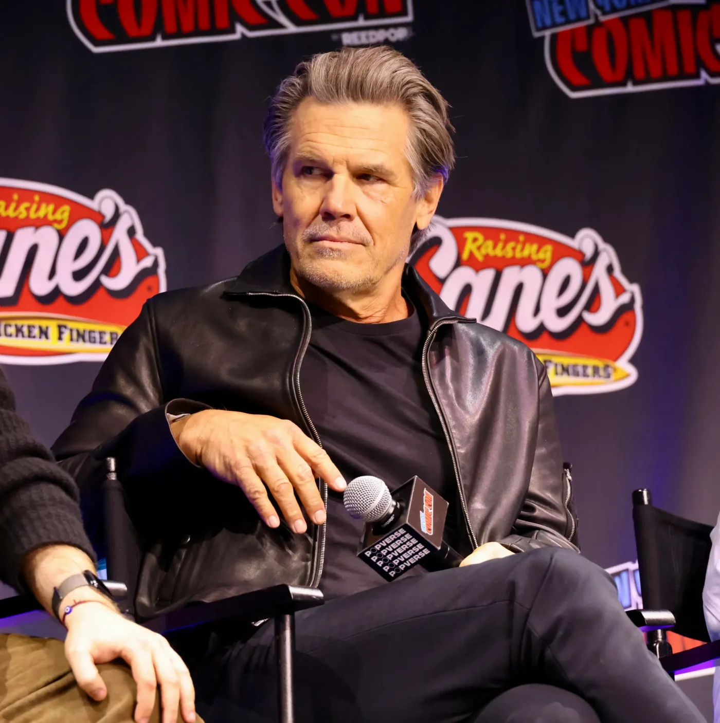 Josh Brolin wearing a black leather jacket and speaking at a panel.