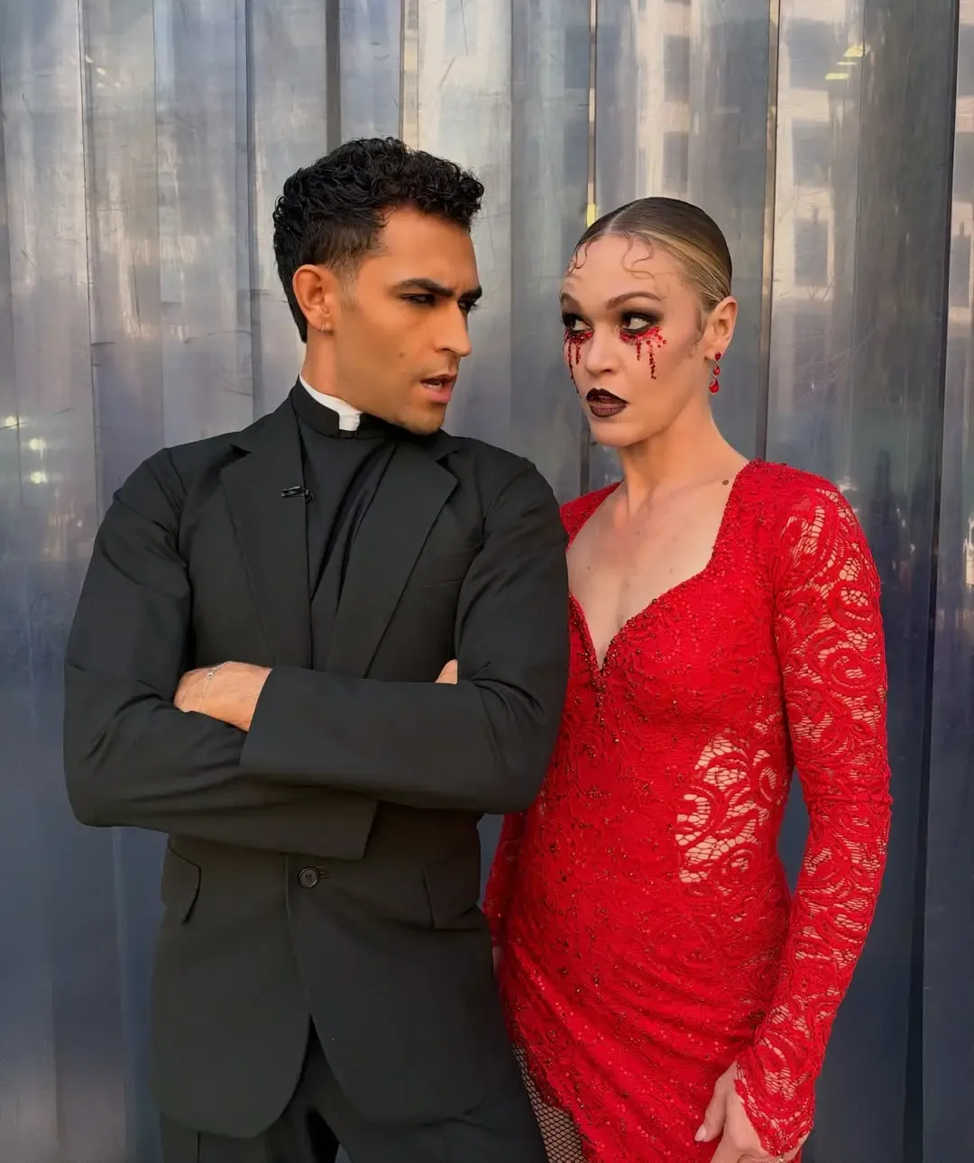 Julia Stiles and Ezra Sosa in costume, with Stiles wearing a red lace dress and dramatic makeup with red tears, and Sosa in a black suit with a clerical collar, glaring at each other during a rehearsal.