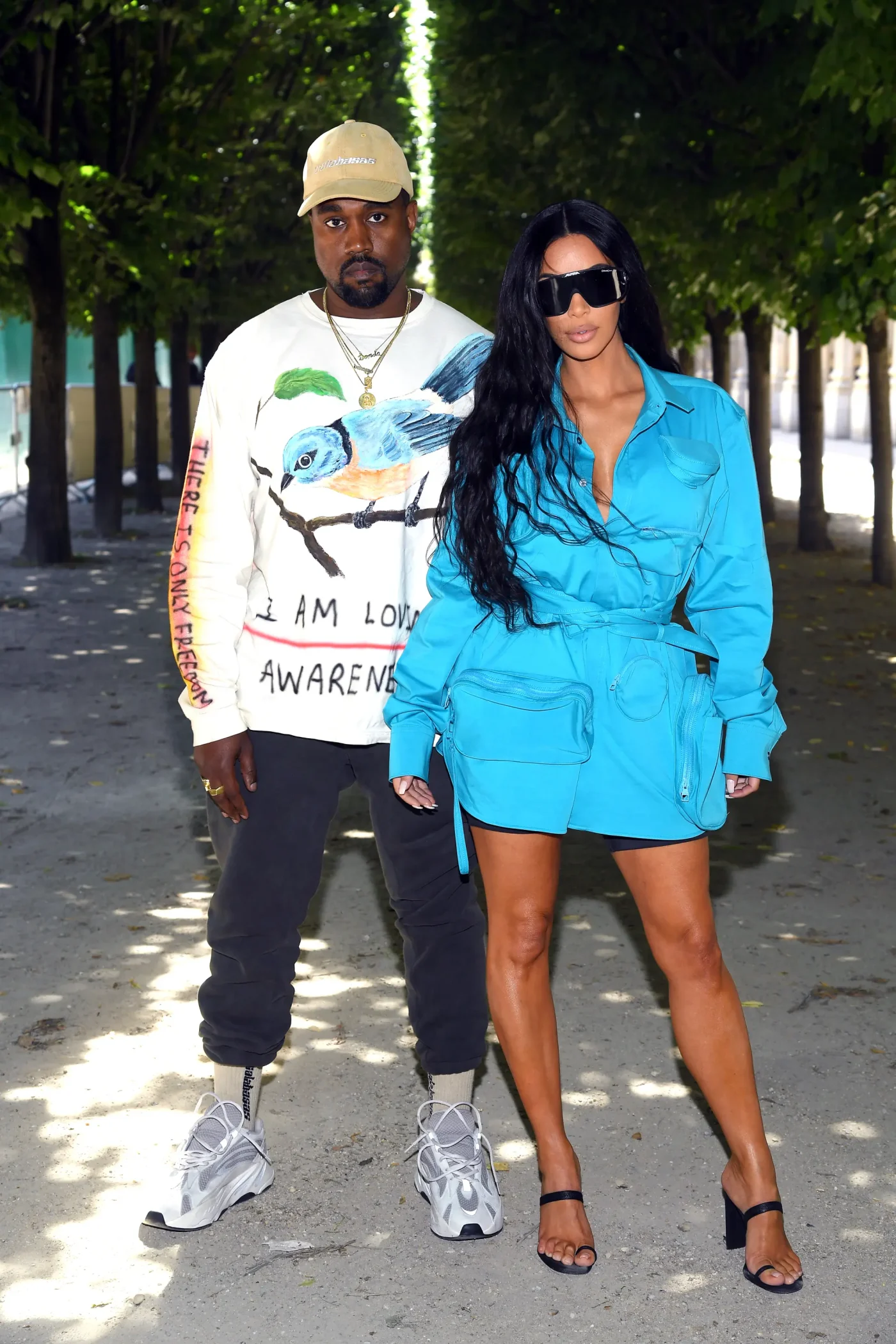Kanye West and Kim Kardashian pose outdoors on a path lined with trees, wearing casual streetwear.