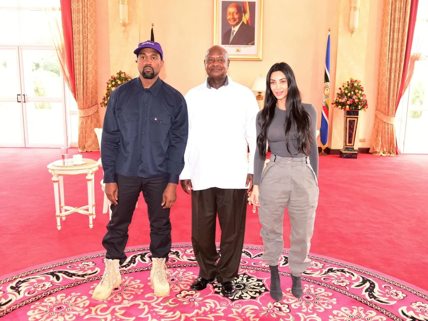 Kanye West, Yoweri Museveni, and Kim Kardashian stand together indoors, with West in a dark blue jacket and purple hat, Museveni in a white shirt and dark trousers, and Kardashian in a gray long-sleeve shirt and cargo pants.