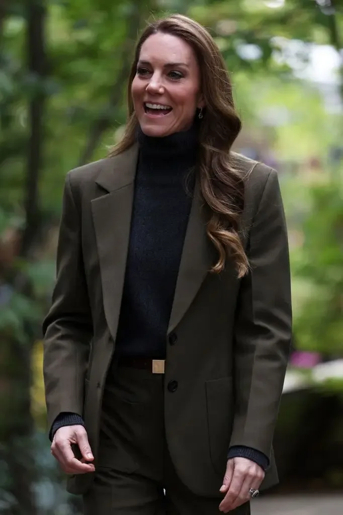 Kate Middleton, wearing a green suit and black turtleneck, smiles while walking outdoors.