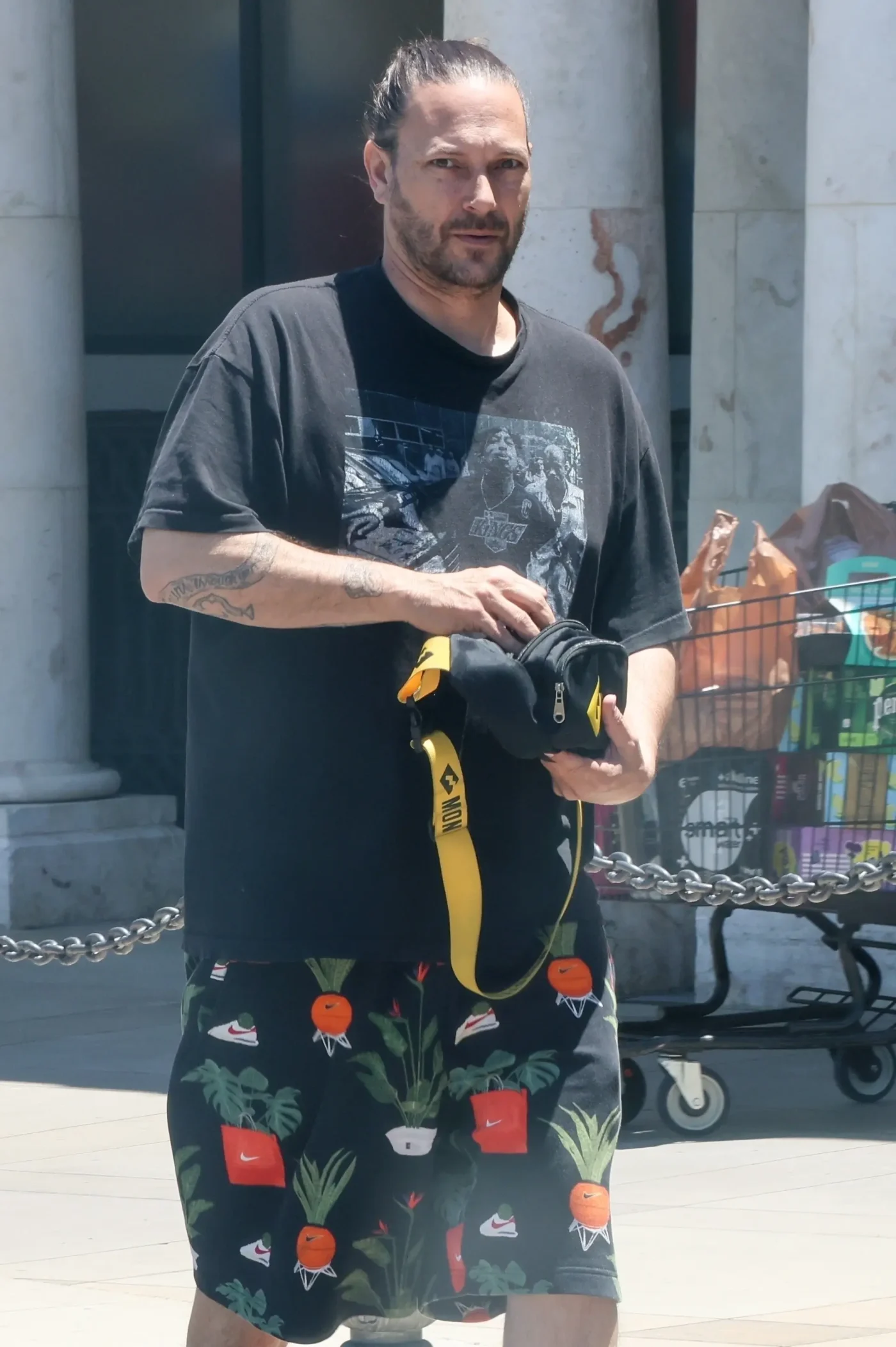 Kevin Federline wearing a black t-shirt and printed shorts while holding a black and yellow bag outside a retail store.