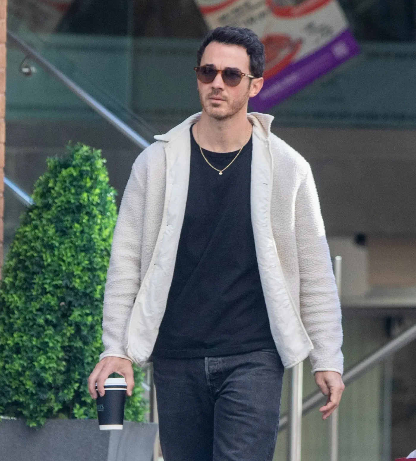 Kevin Jonas wearing sunglasses, a black t-shirt, and a cream-colored fleece jacket, holding a coffee cup while walking in Toronto.