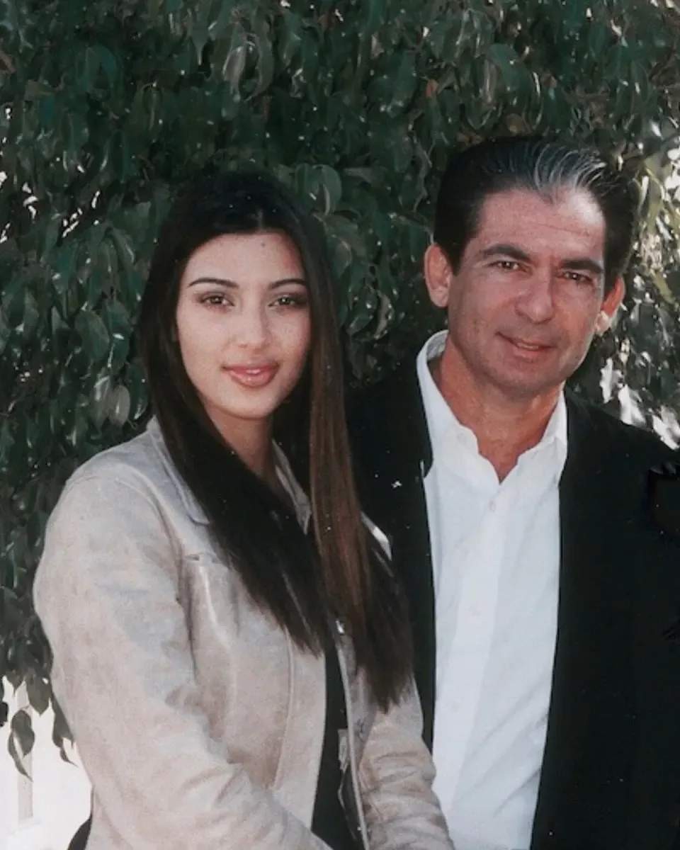 Kim Kardashian and her father, Robert Kardashian Sr., standing together in front of leafy foliage.