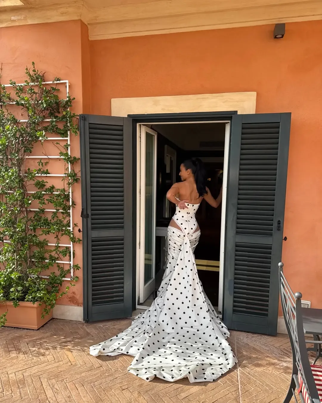 Kim Kardashian in a white and black polka dot dress with a long train, standing in a doorway against an orange wall.