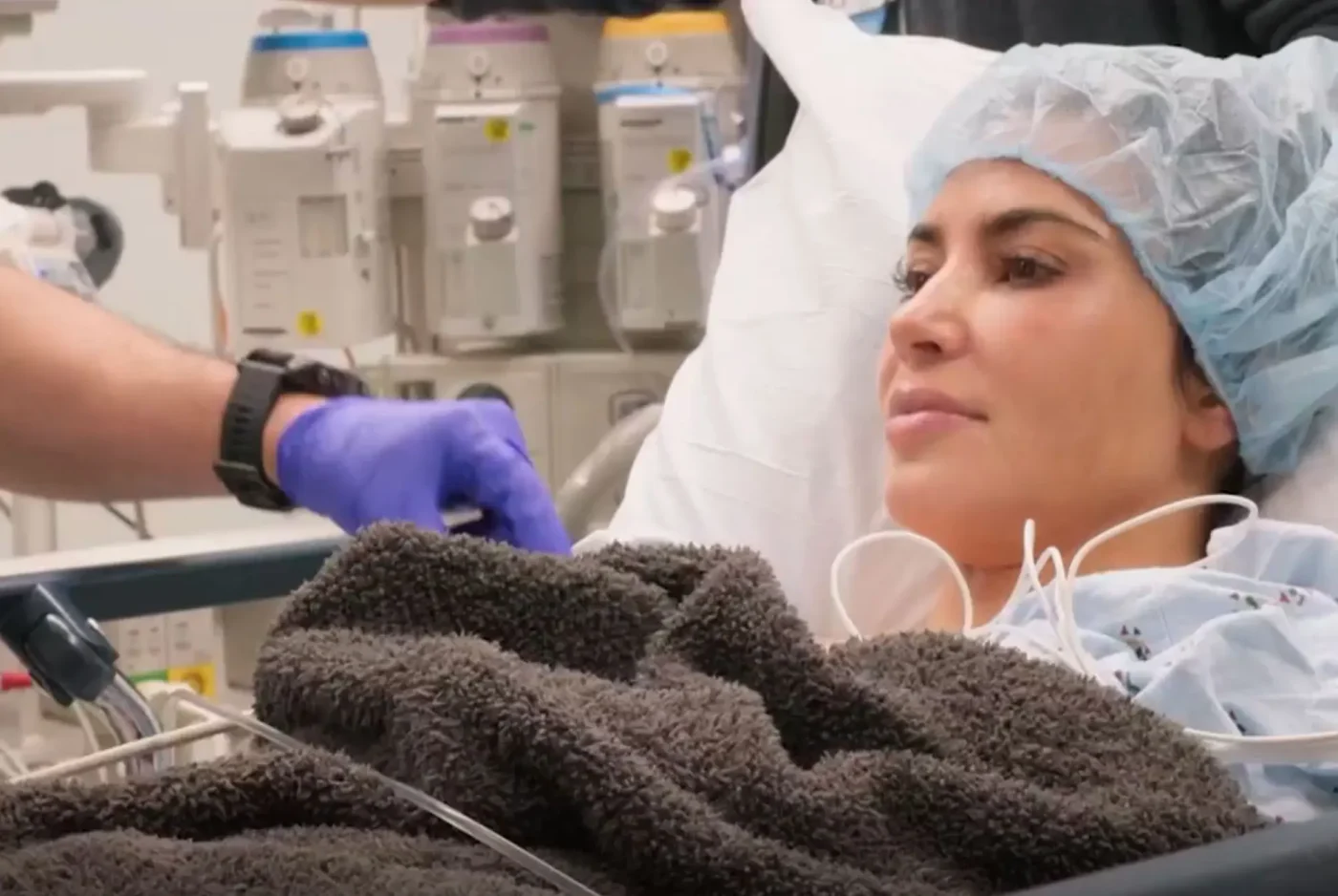 Kim Kardashian lies in a hospital bed wearing a hairnet, with medical equipment in the background.