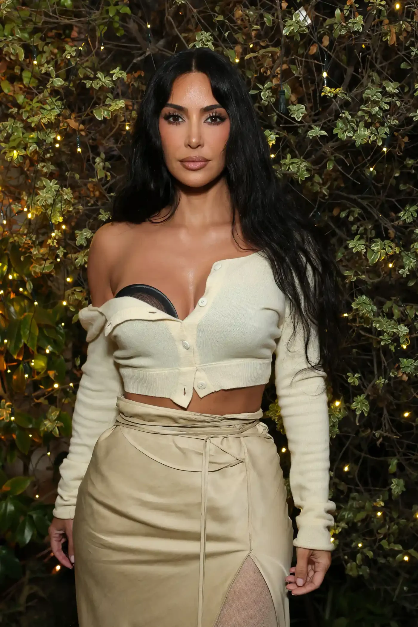 Kim Kardashian poses outside in a paved garden in front of a wall covered in vines and yellow fairy lights. she is wearing a satin skirt with a gauzy thigh-high slit and an off-the-shoulder cardigan in cream over a see-through black bra.