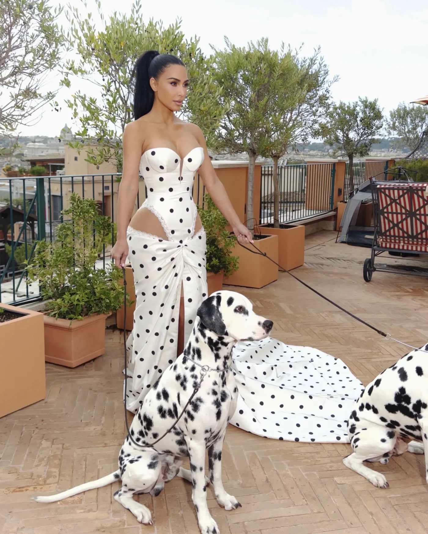 Kim Kardashian wearing a white polka-dot dress on a terrace, holding leashes of two dalmatians.