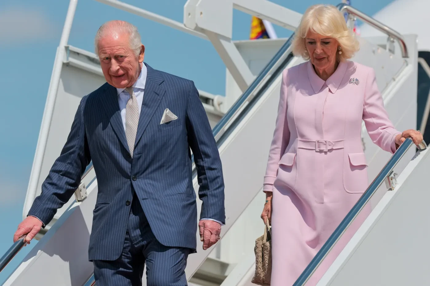 King Charles and Queen Camilla disembark a plane. Charles is looking at the camera and wearing a dark blue pinstriped suit as he holds onto the railing. Camilla is wearing a light pink trench coat and has her eyes downcast as she holds onto the railing.