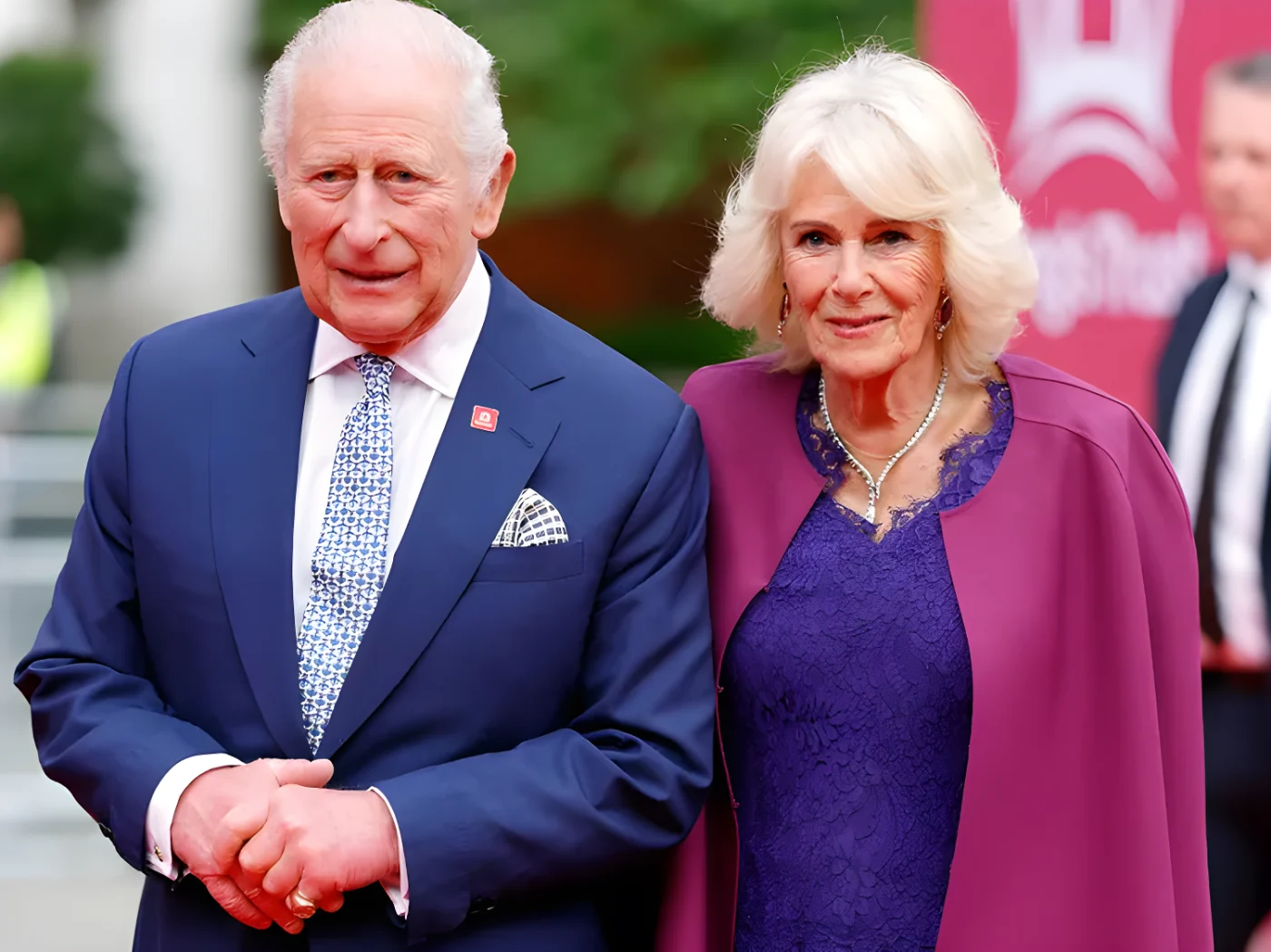 King Charles and Queen Camilla walk side by side. CHarles is wearing a dark blue suit over a white dress shirt and light blue patterned tie. Camilla is wearing a purple lace evening gown beneath a pink cape jacket.
