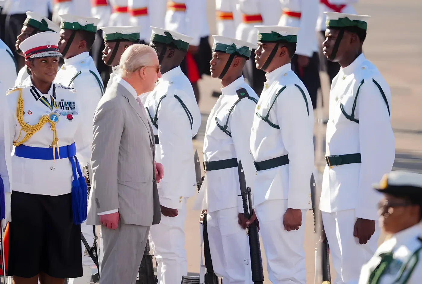 King Charles in a grey suit and pink tie stands with military officers in white uniforms.