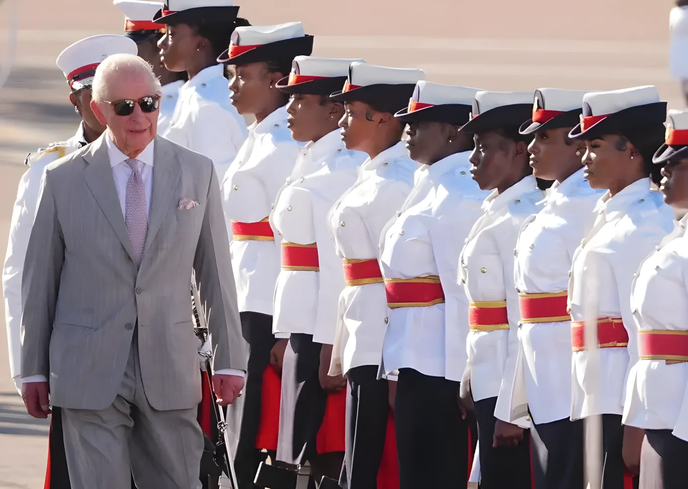 King Charles in a grey suit and sunglasses, walks past a line of uniformed women.