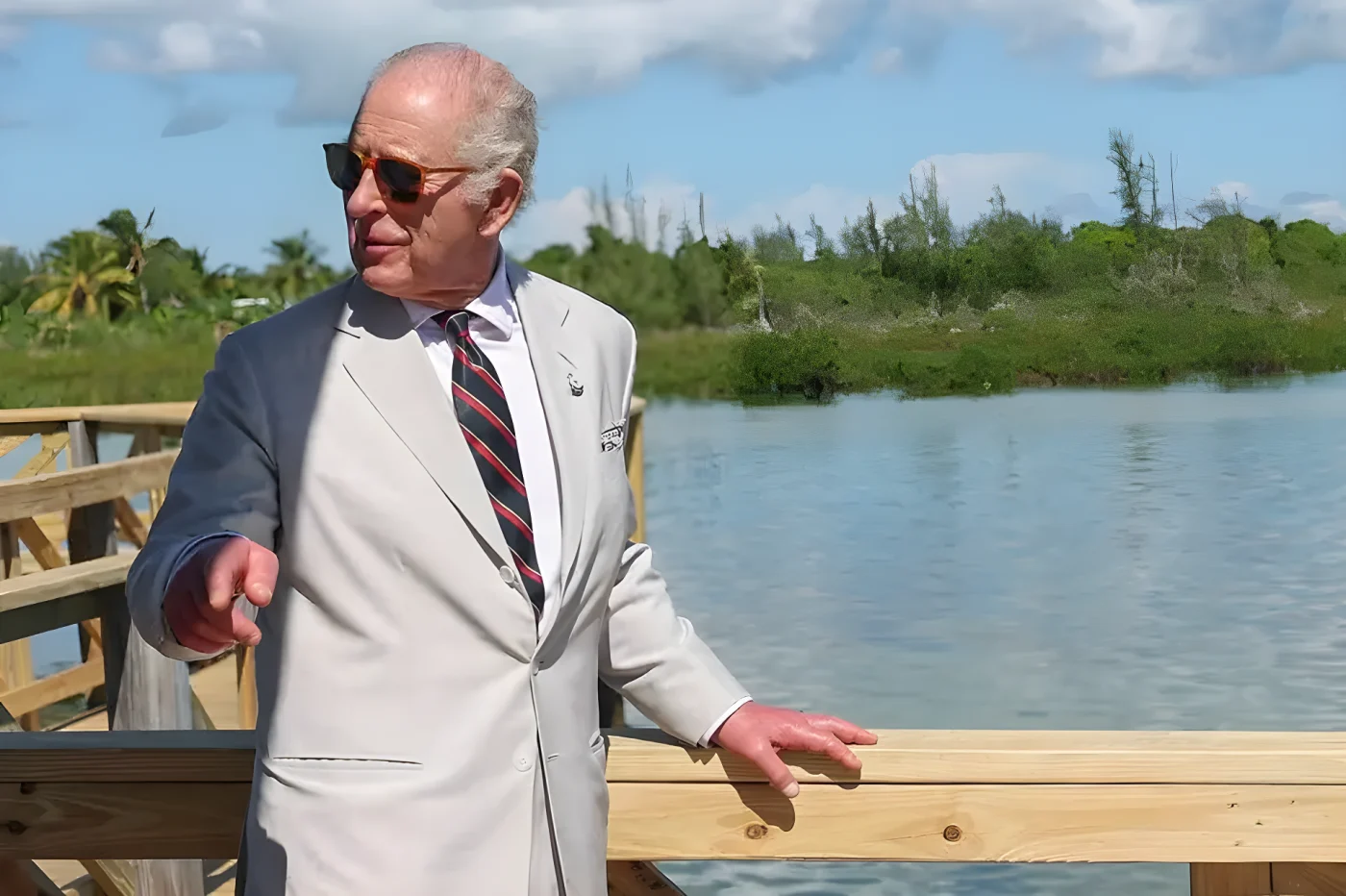 King Charles in a light suit and striped tie, stands by a wooden dock by a lake.