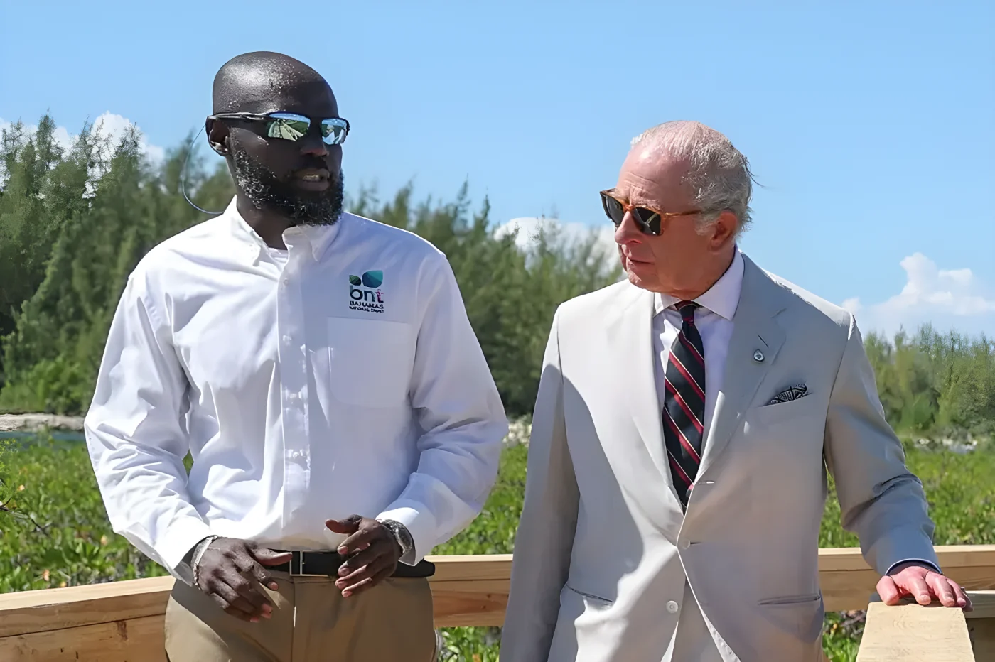 King Charles in a light suit and striped tie, talks with a man in a white shirt and sunglasses.
