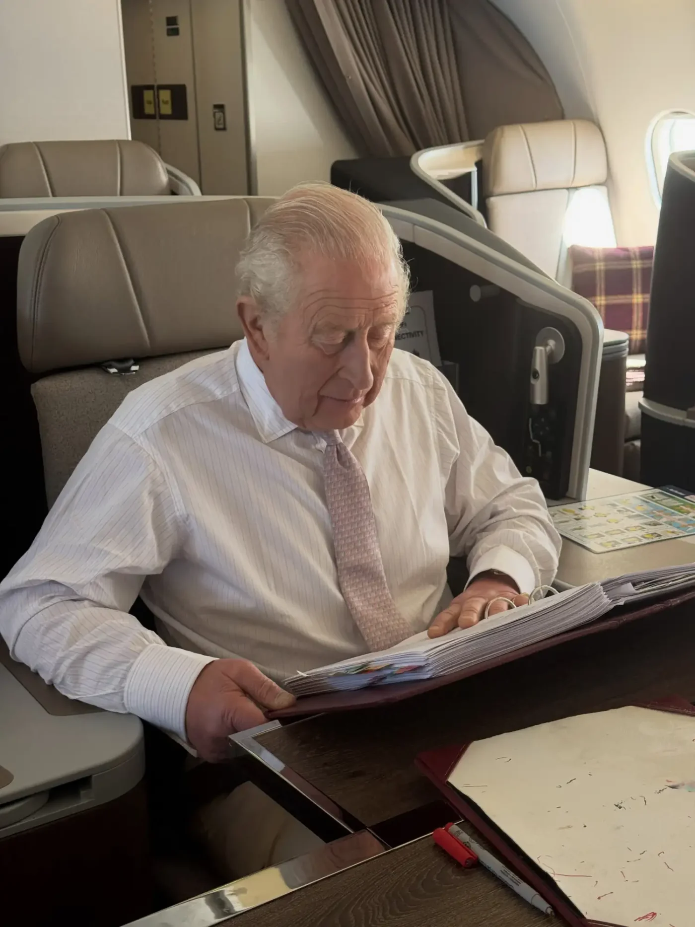King Charles reads a binder of briefing papers as he sits aboard a private plane. He is wearing a white dress shirt and a light pink tie.