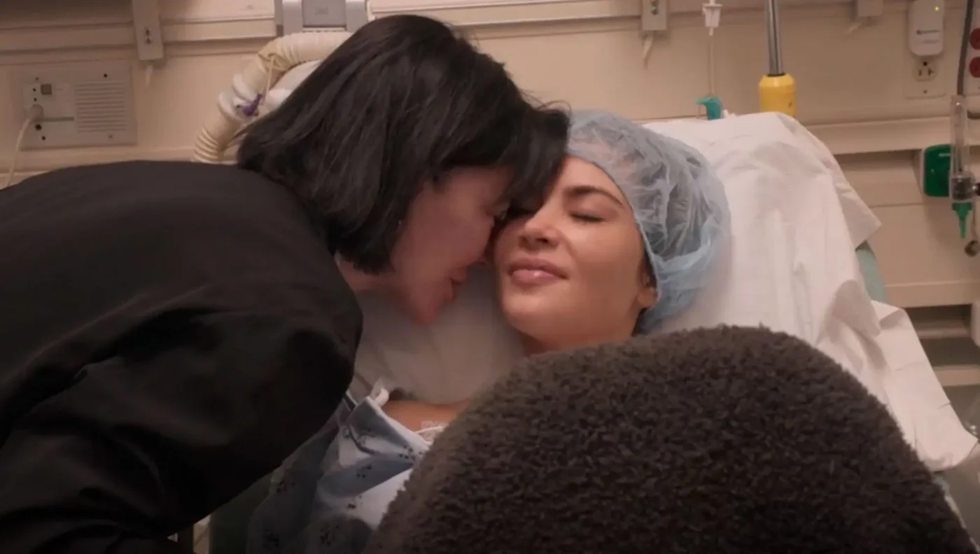Kris Jenner leans over to kiss Kim Kardashian, who is lying in a hospital bed wearing a hairnet.
