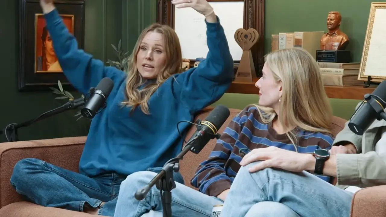 Kristen Bell gestures wildly while speaking into a microphone on Armchair Expert, with Monica Padman sitting beside her.