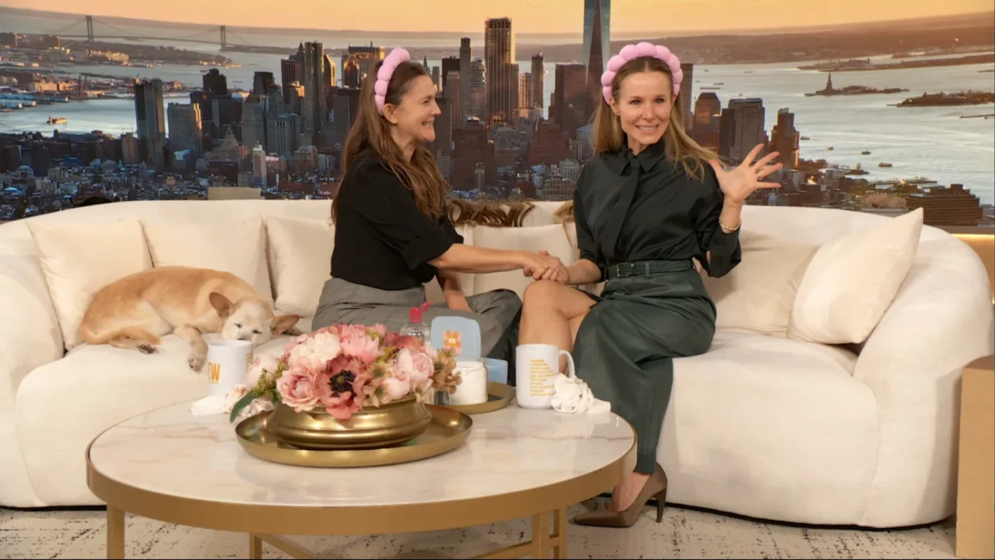 Kristen Bell sitting on a sofa with another person both wearing pink headbands a cityscape visible in the background