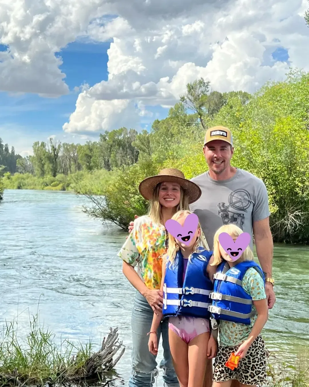 Kristin Bell and Dax Shepard pose outdoors with their two daughters by a river on a sunny day, with the daughters