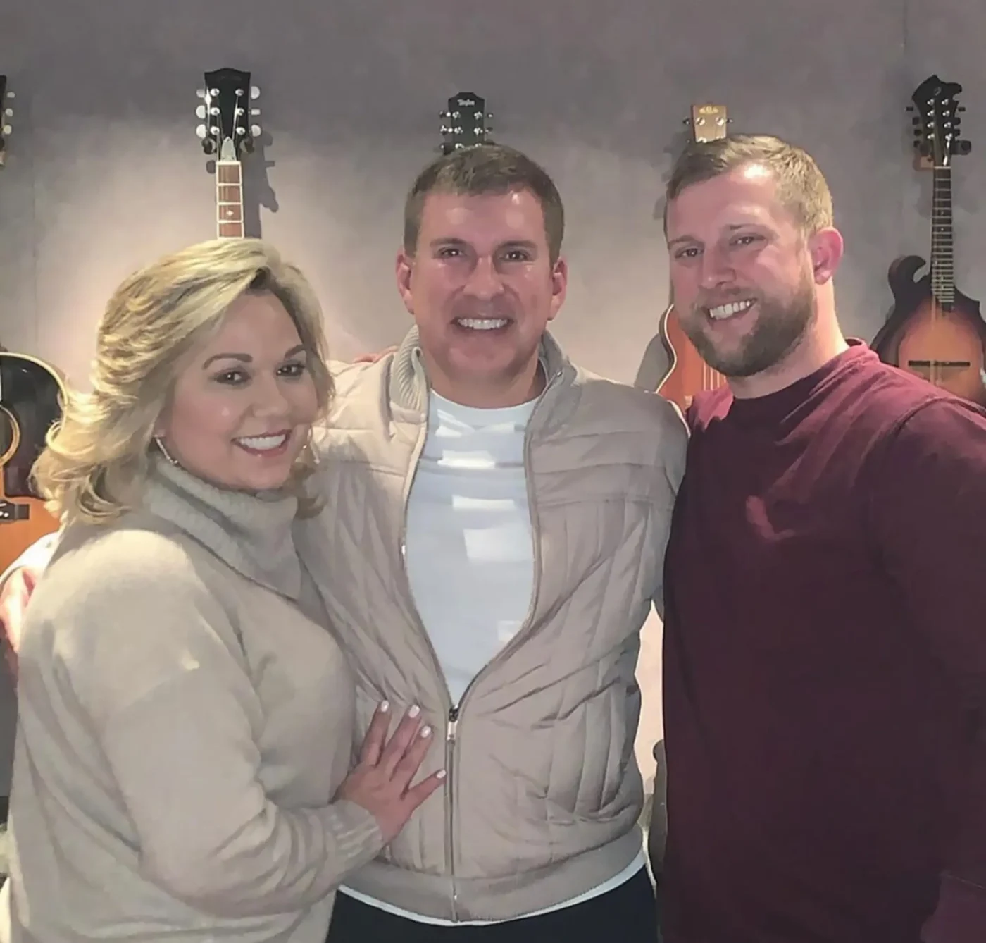 Kyle Chrisley smiling alongside Todd and Julie Chrisley, with guitars hanging on the wall behind them.