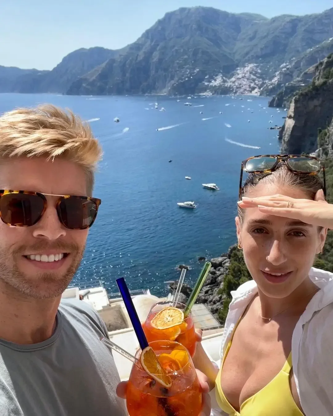 Kyle Cooke and Amanda Batula holding drinks on a balcony overlooking the sea.