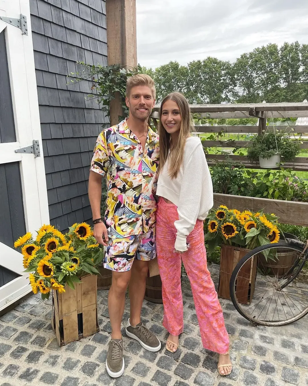 Kyle Cooke and Amanda Batula standing outside near wooden crates filled with sunflowers.